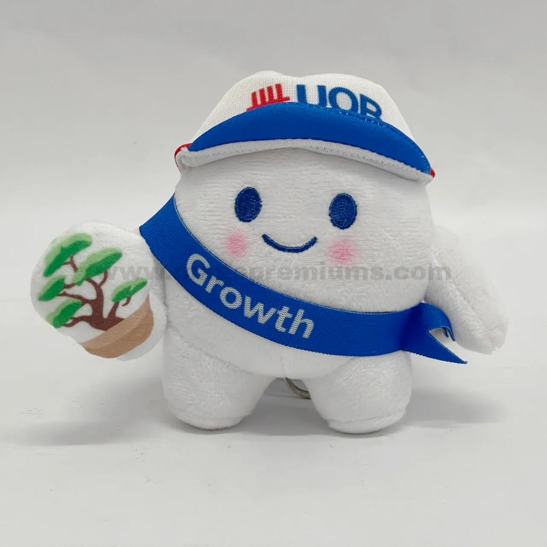 Plush Keychain Custom plushy soft toy mascot with embroidered smile, branded cap, blue sash, and tree accessory, designed as a promotional plush keychain for corporate branding, events, and marketing campaigns.
